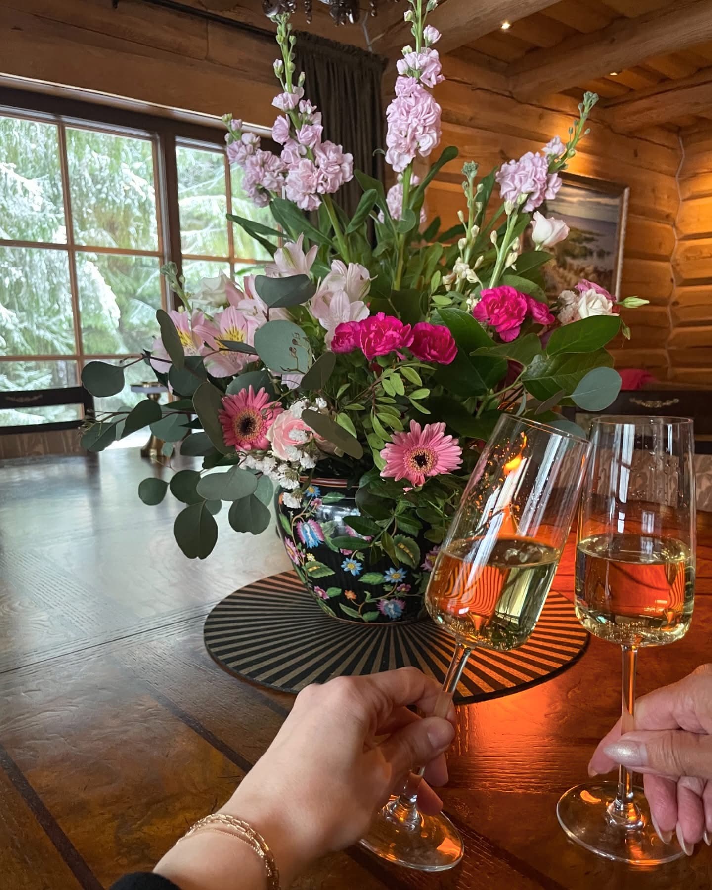 Two glasses raised beside a tall arrangement of flowers, snow through the window