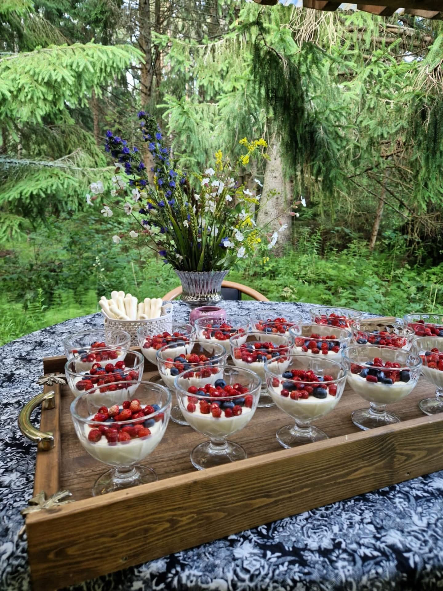 Berry desserts in coupe glasses on a wooden tray, wildflowers and spruce behind