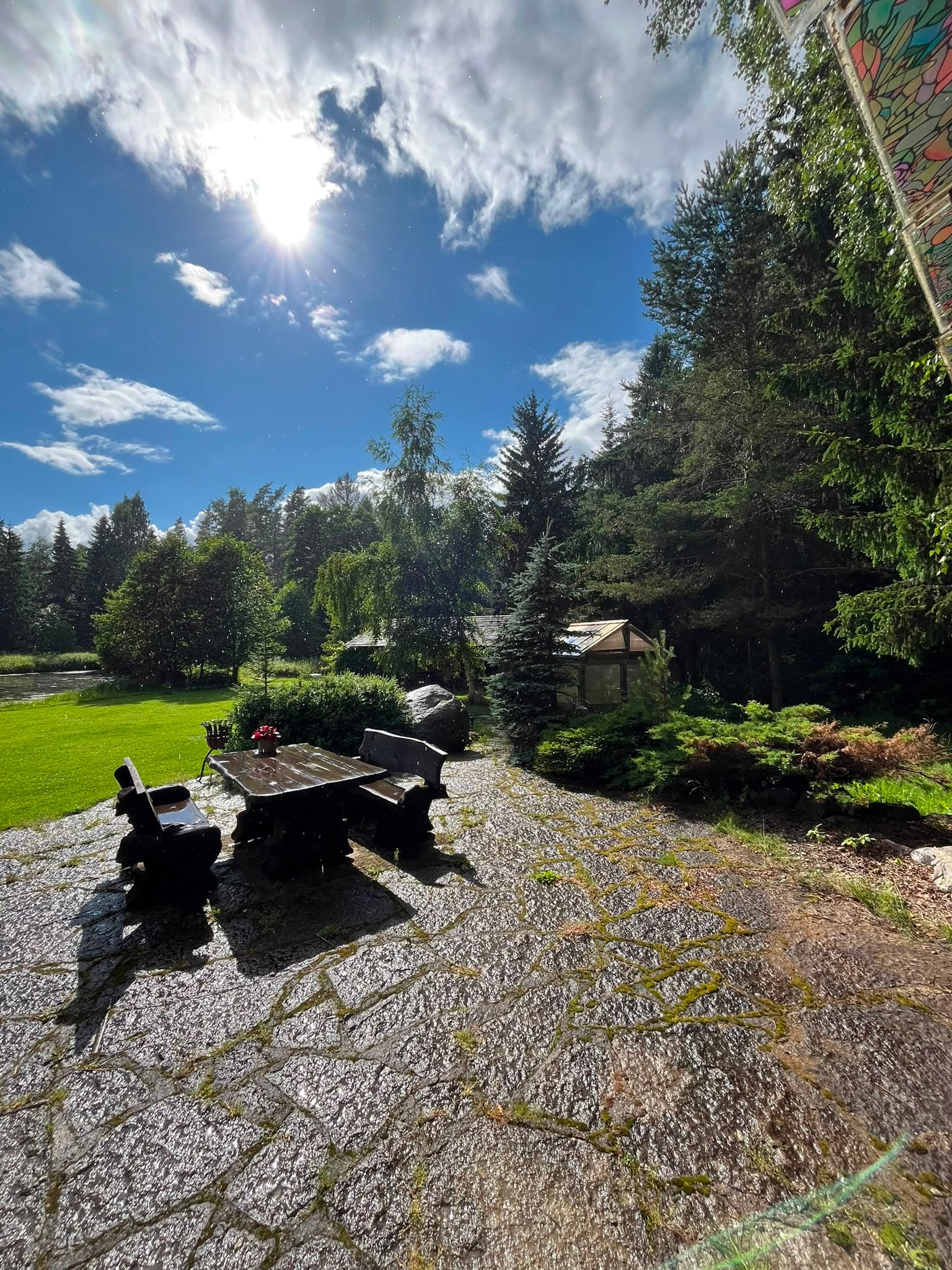 A dark timber table and benches on the flagstone terrace, spruces and the greenhouse beyond